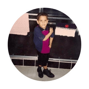 A baby photo of Mark, in a bathroom, posing with his arm on his hip and his other arm on his shoulder with his legs crossed and inverted. He has short black hair and is wearing black shorts, a navy and pink shirt, and black shoes.