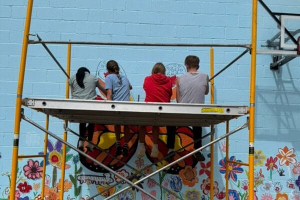 Students sit on an elevated structure while painting a mural on a large outdoor building.